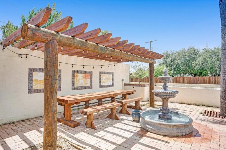 A wooden bench sitting under an arbor near a fountain.